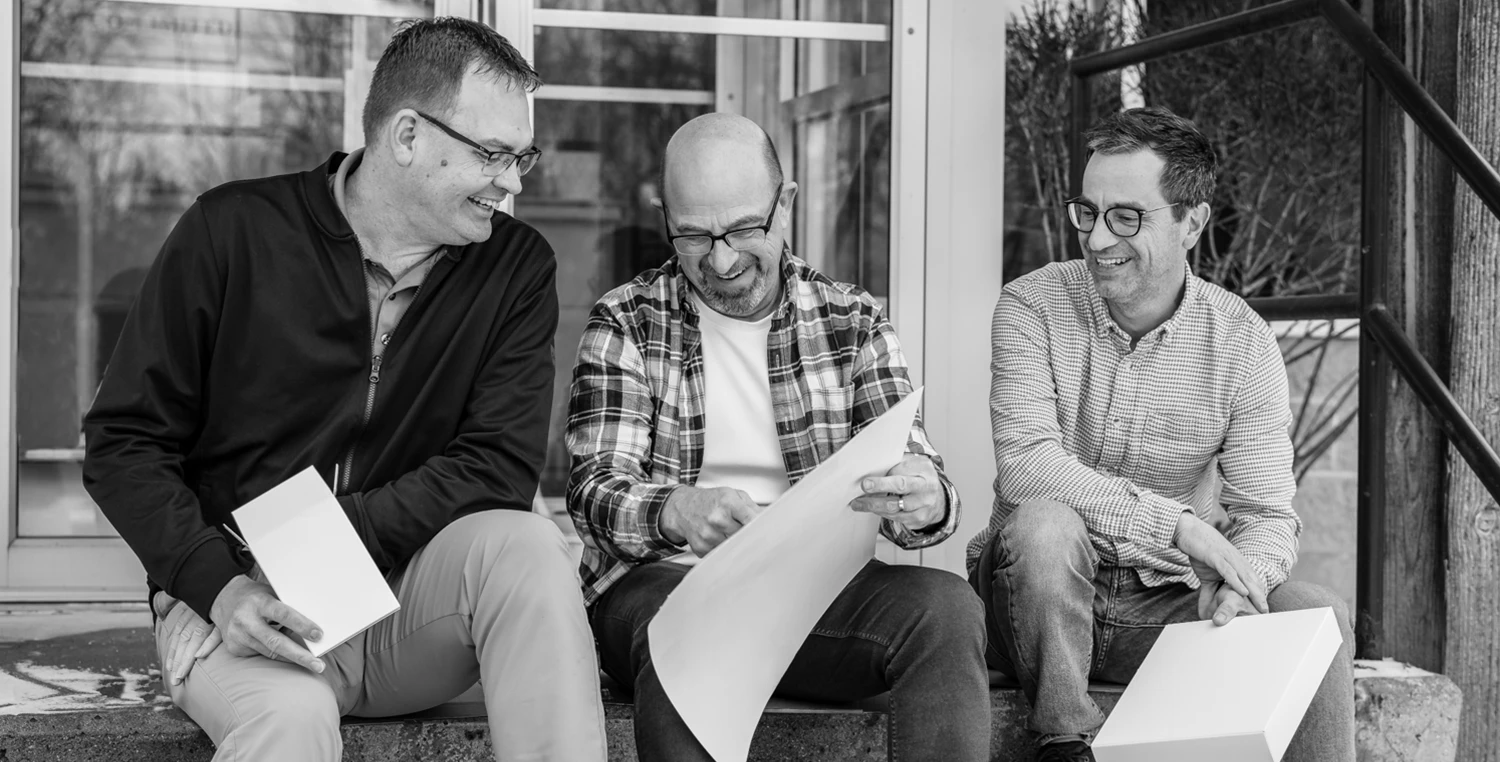 Three members of the Ingersoll Paper Box team casually seated on steps outside their facility, smiling and examining a prototype carton.